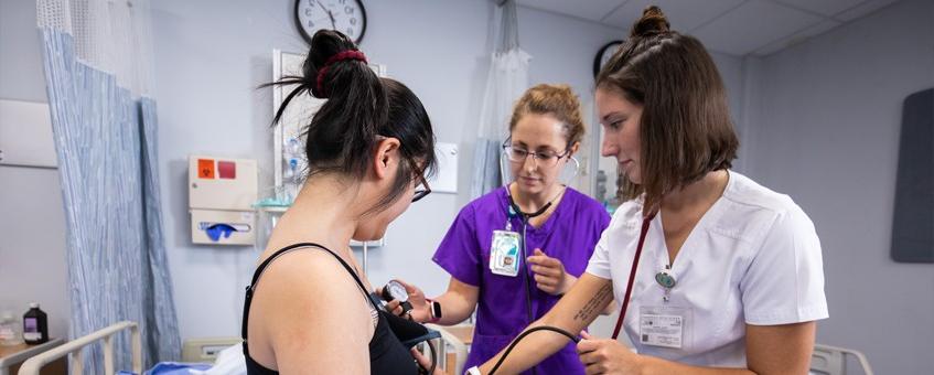 A nursing instructor demonstrates a medical principle on a volunteer alongside a student.